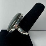 Silver ring with a large oval labradorite stone featuring subtle blue and green iridescence, displayed on a black velvet ring holder against a light background.