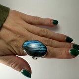 Hand wearing an oval labradorite ring displaying iridescent blue and green hues, with matching dark green nail polish on fingers gently spread.