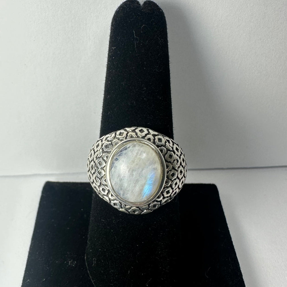 Oval moonstone set in a wide silver band with a geometric hexagonal pattern, displayed on a black velvet ring stand against a neutral background.