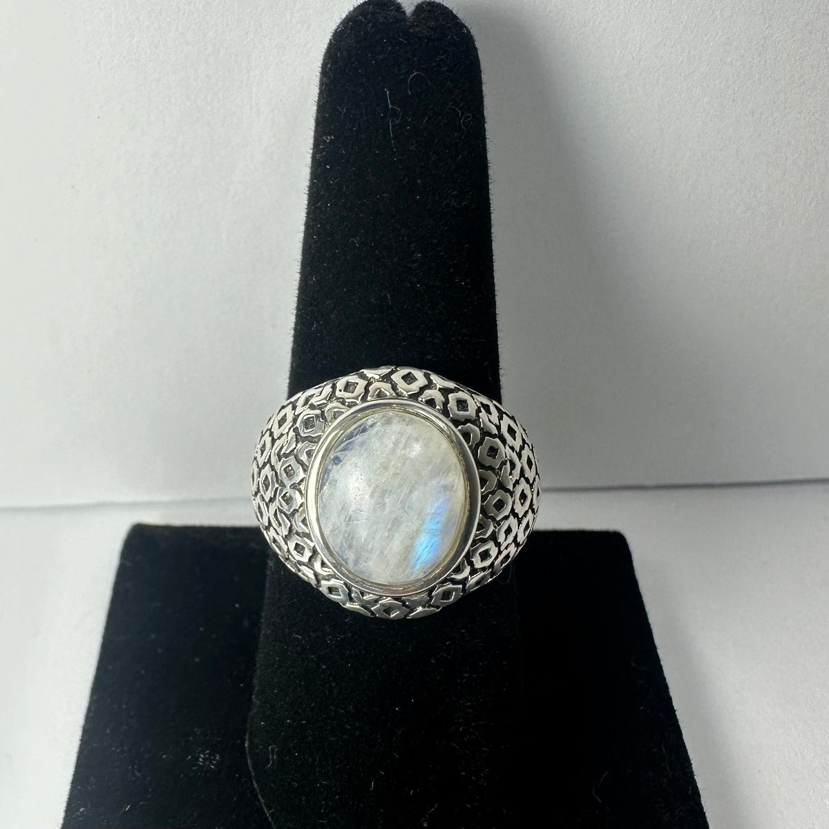 Oval moonstone set in a wide silver band with a geometric hexagonal pattern, displayed on a black velvet ring stand against a neutral background.