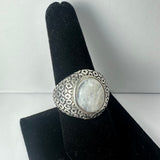 Silver ring with an oval moonstone centerpiece set in an intricately patterned band, displayed on a black velvet finger stand against a plain white background.