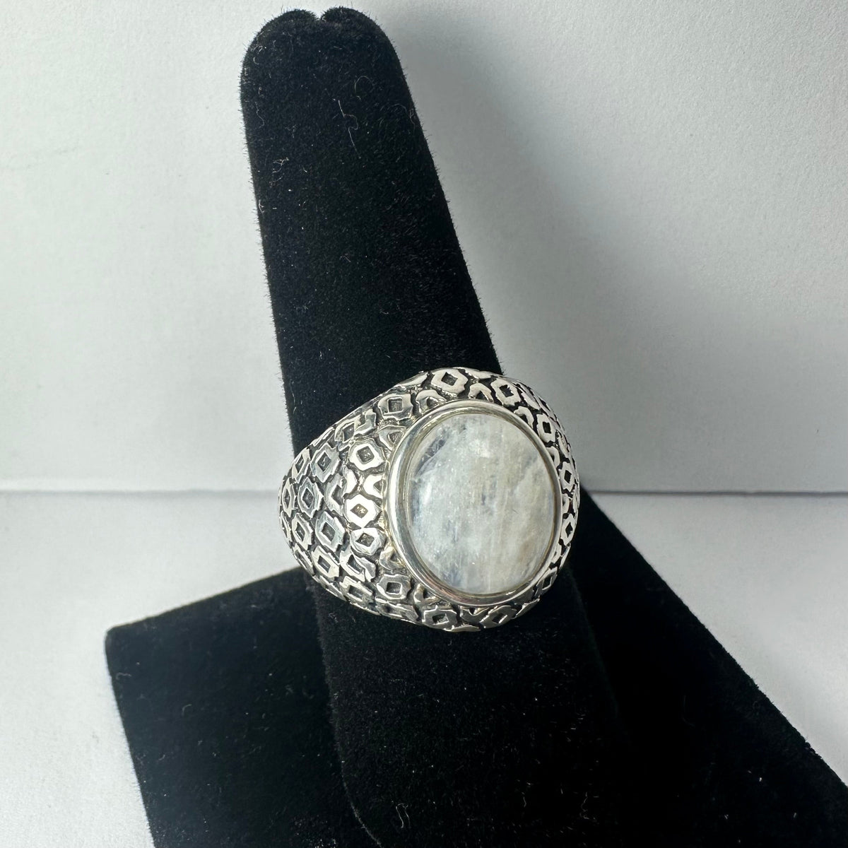 Silver ring with an oval moonstone centerpiece set in an intricately patterned band, displayed on a black velvet finger stand against a plain white background.