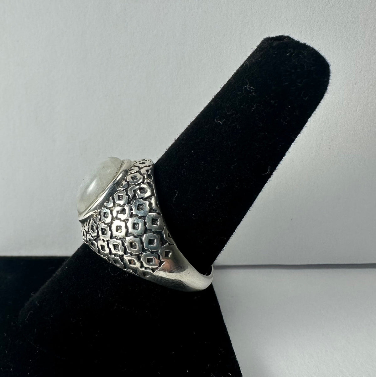 Silver ring with a rounded moonstone cabochon on top, featuring a textured geometric pattern on the band, displayed on a black velvet ring holder.