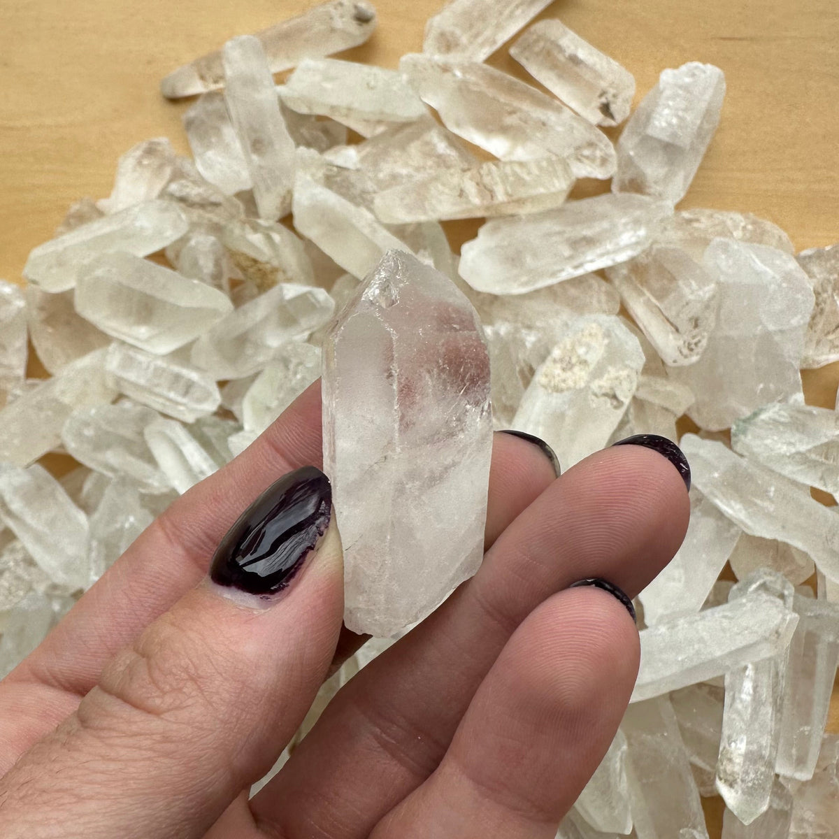 Hand holding a pointed translucent Brazilian quartz crystal with a cluster of similar clear quartz crystals in the background on a wooden surface.