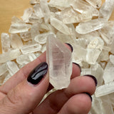 Hand holding a clear, pointed Brazilian quartz crystal against a background of multiple similar clear quartz points on a wooden surface.