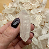 Hand holding a clear, pointed quartz crystal with natural inclusions above a pile of similar clear, hexagonal quartz crystal points on a wooden surface.