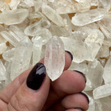 Close-up of a hand with dark nail polish holding a clear, pointed Brazilian quartz crystal in front of a pile of similar translucent crystals.