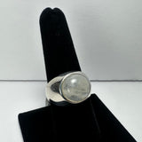 Silver ring with a large round moonstone displaying translucent white and gray hues, shown on a black velvet finger display against a plain white background.