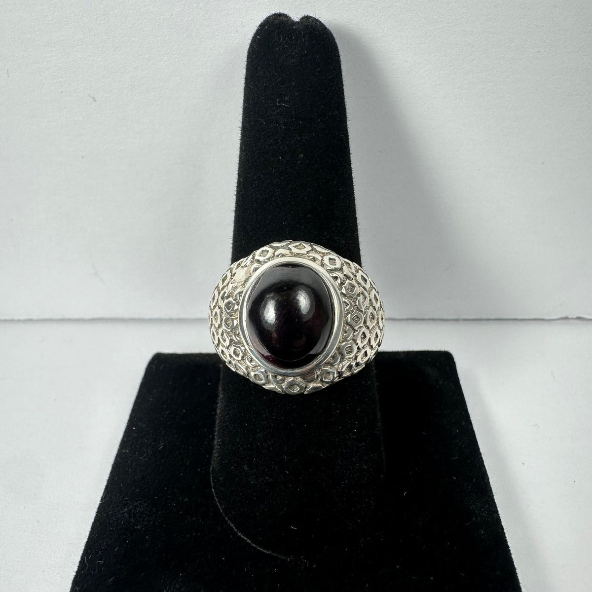 Silver ring with a large oval garnet cabochon set in a textured band, displayed upright on a black velvet ring holder against a plain white background.