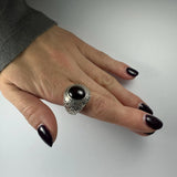 Hand with dark polished nails wearing a silver ring featuring an oval red garnet center stone and textured band on the middle finger