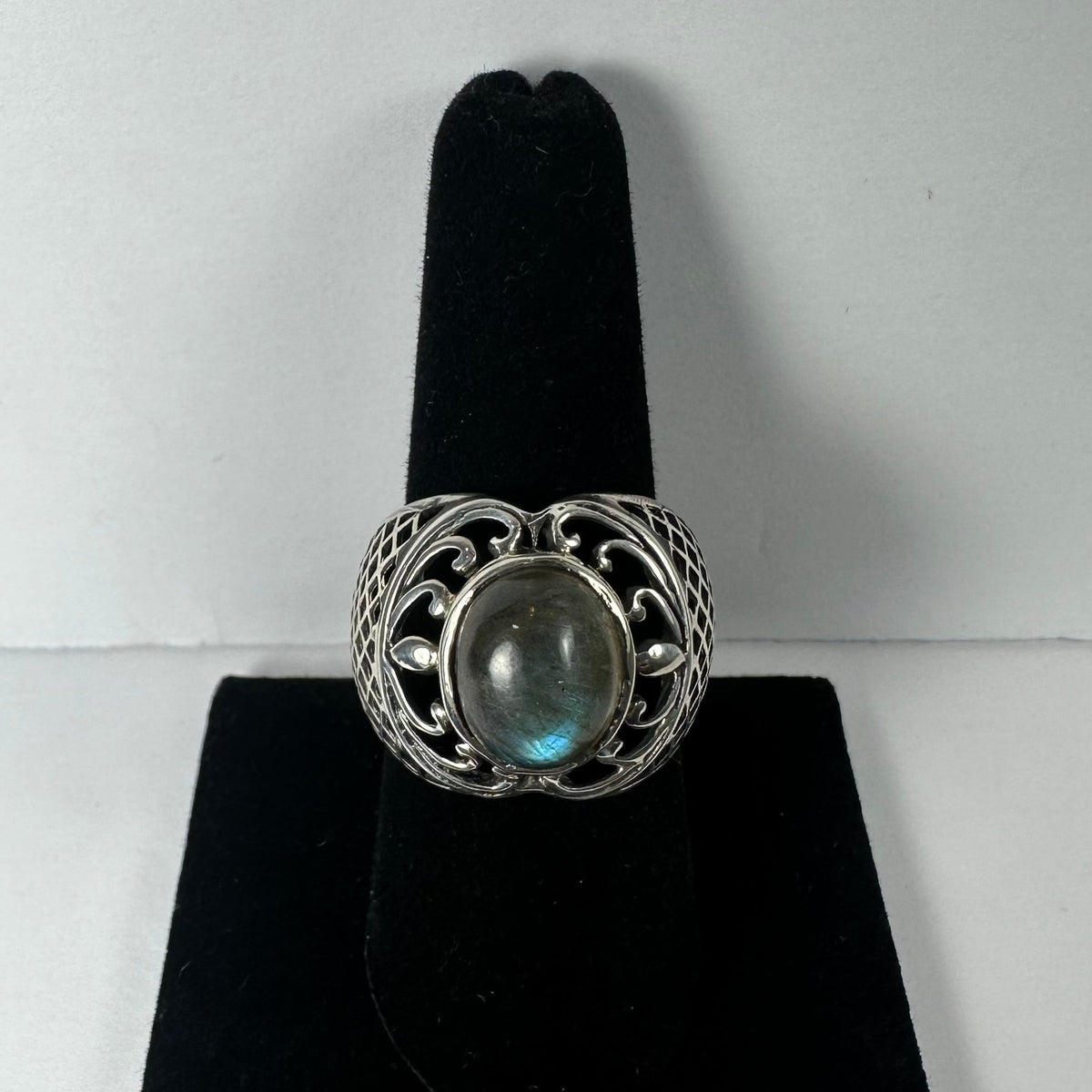 Silver ring with intricate openwork design featuring a central oval labradorite stone showing subtle blue and green flashes, displayed on a black ring holder.
