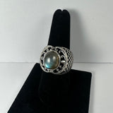 Silver ring with a large oval labradorite stone featuring gray and blue flashes, set in an intricate openwork band with a lattice pattern on the sides, displayed on a black velvet stand.