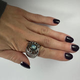 Silver ring with an oval labradorite stone showing blue and green flashes, featuring intricate metalwork around the bezel and textured band, worn on a hand with dark nail polish.