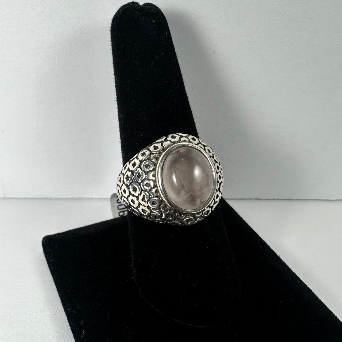 Silver ring with a polished round rose quartz stone set in an intricately patterned band, displayed on a black velvet ring holder against a white background.