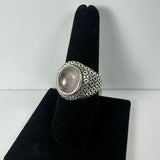 Silver ring with a large round rose quartz cabochon set in an intricately patterned band, displayed on a black velvet ring holder against a white background.