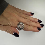 Hand with black polished nails wearing a silver ring featuring a smooth, oval rose quartz stone set in an intricately patterned band on the ring finger.