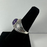Silver ring with textured geometric pattern and a domed purple amethyst stone, displayed on a black velvet ring holder against a light gray background.