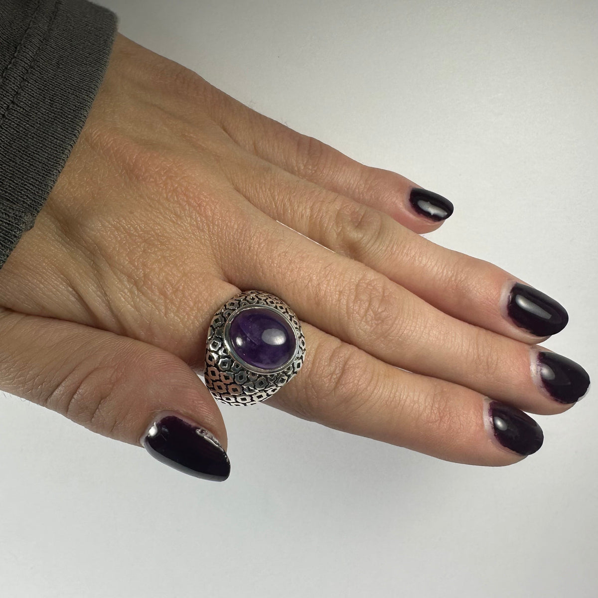 Hand with dark purple painted nails wearing a silver ring featuring a polished oval amethyst stone in the center, shown against a plain white background.