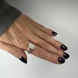 Hand with dark purple polished nails wearing a silver ring featuring a round translucent moonstone on the middle finger against a plain white background.
