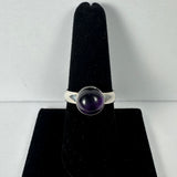 Silver ring with a round, smooth amethyst cabochon set on a simple band, displayed upright on a black velvet ring holder against a white background.