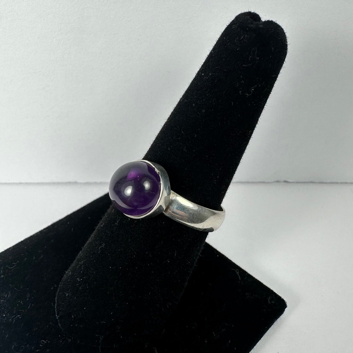 Silver ring featuring a polished, rounded amethyst stone set in a simple bezel on a black velvet ring display stand.