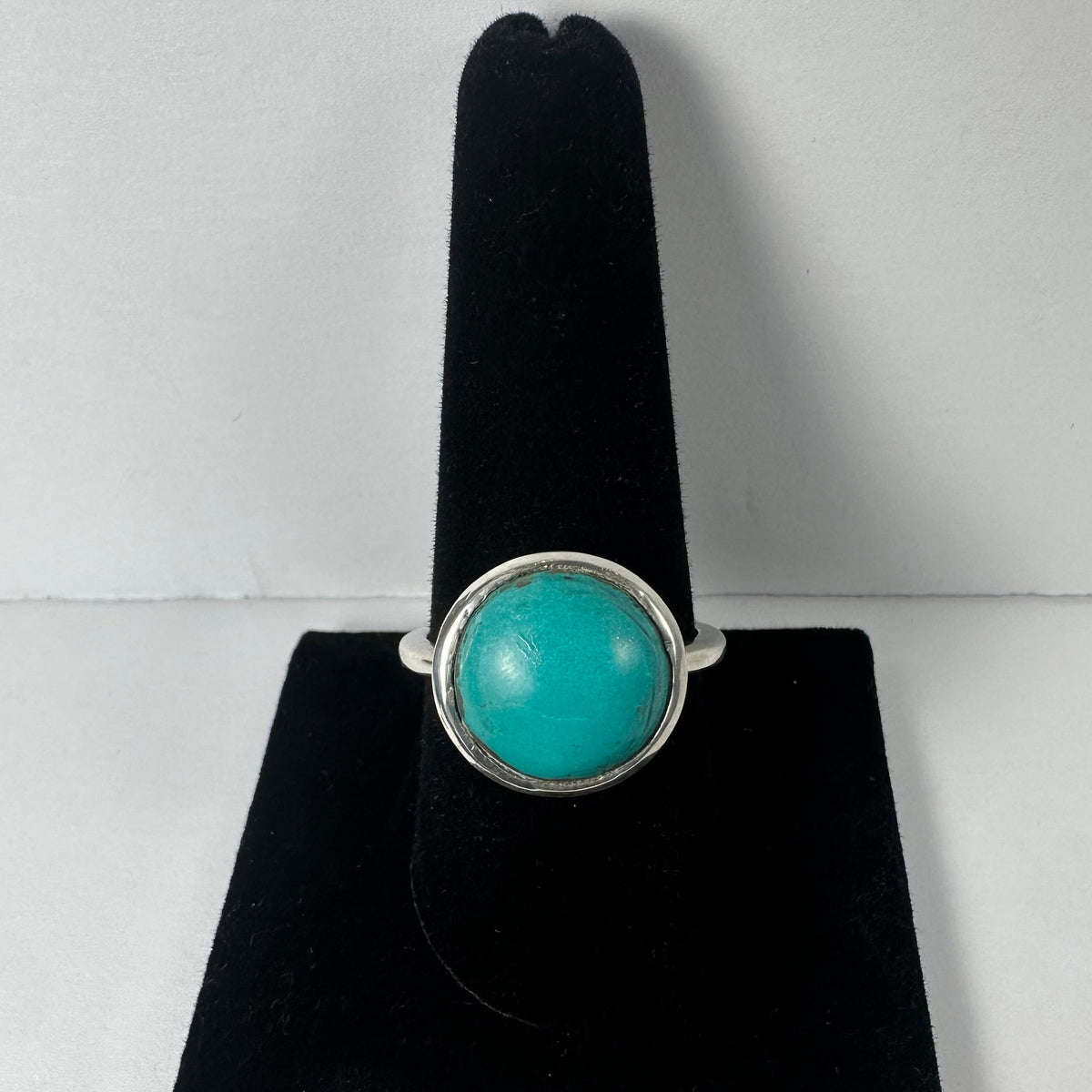 Silver ring with a smooth, round turquoise stone set in a simple bezel, displayed upright on a black velvet ring holder against a white background.