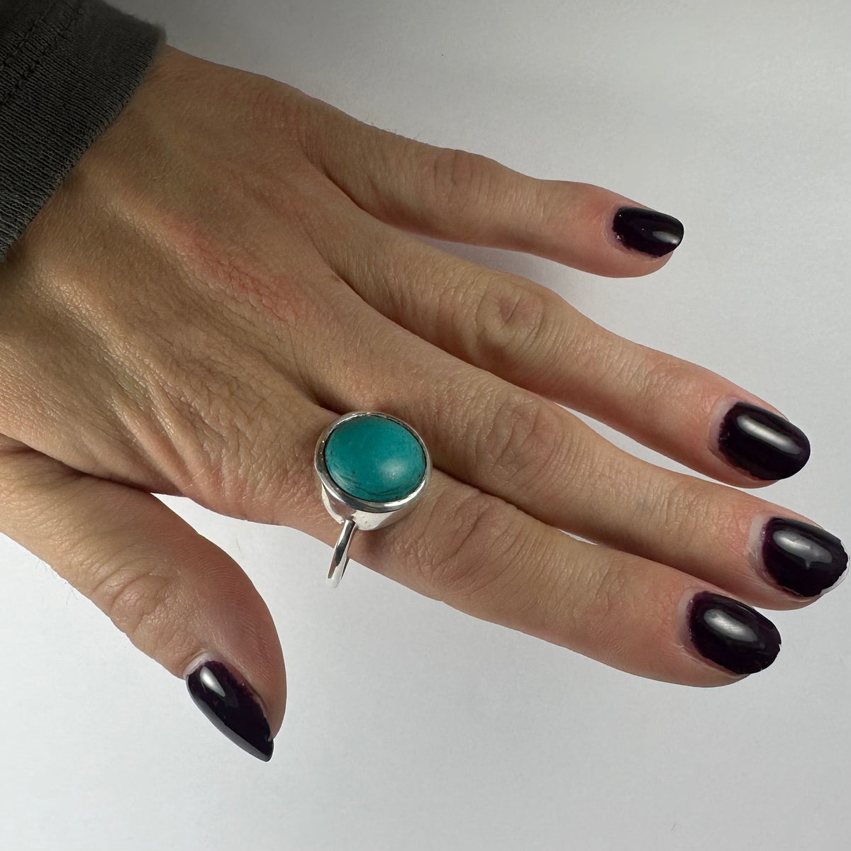 Hand with dark purple polished nails wearing a silver ring featuring a round, bright turquoise stone on the ring finger against a plain white background.