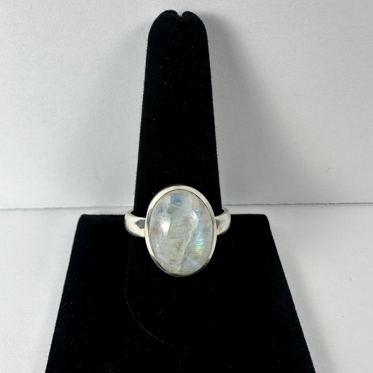 Silver ring with a polished oval moonstone exhibiting subtle blue and rainbow flashes, displayed upright on a black velvet stand against a plain white background.