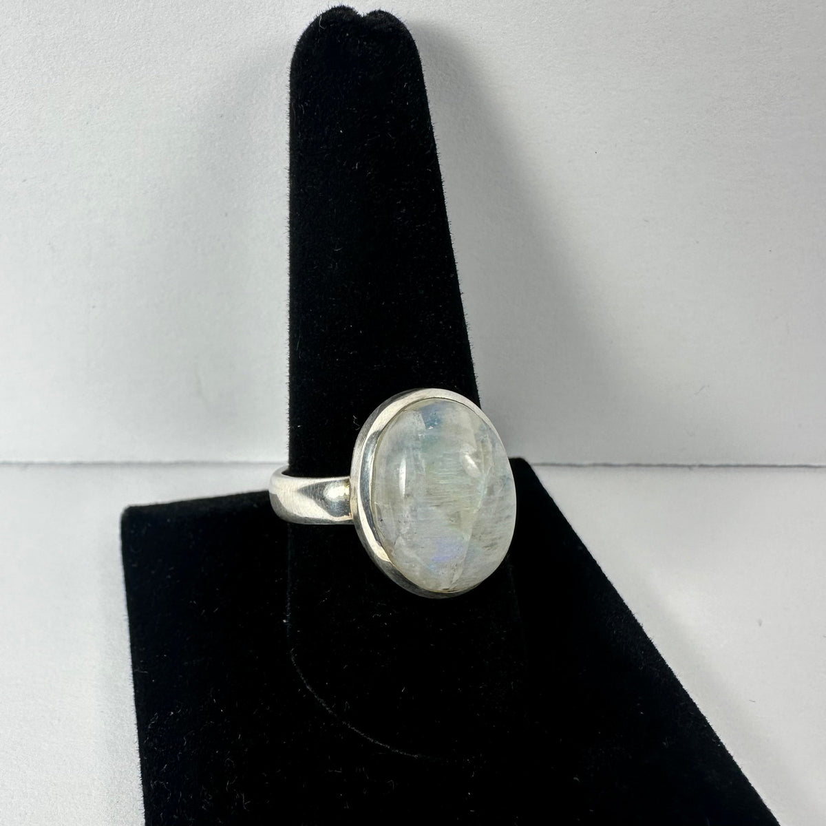 Silver ring with a polished oval moonstone displaying subtle blue and white reflections, set on a black velvet ring holder against a plain white background.