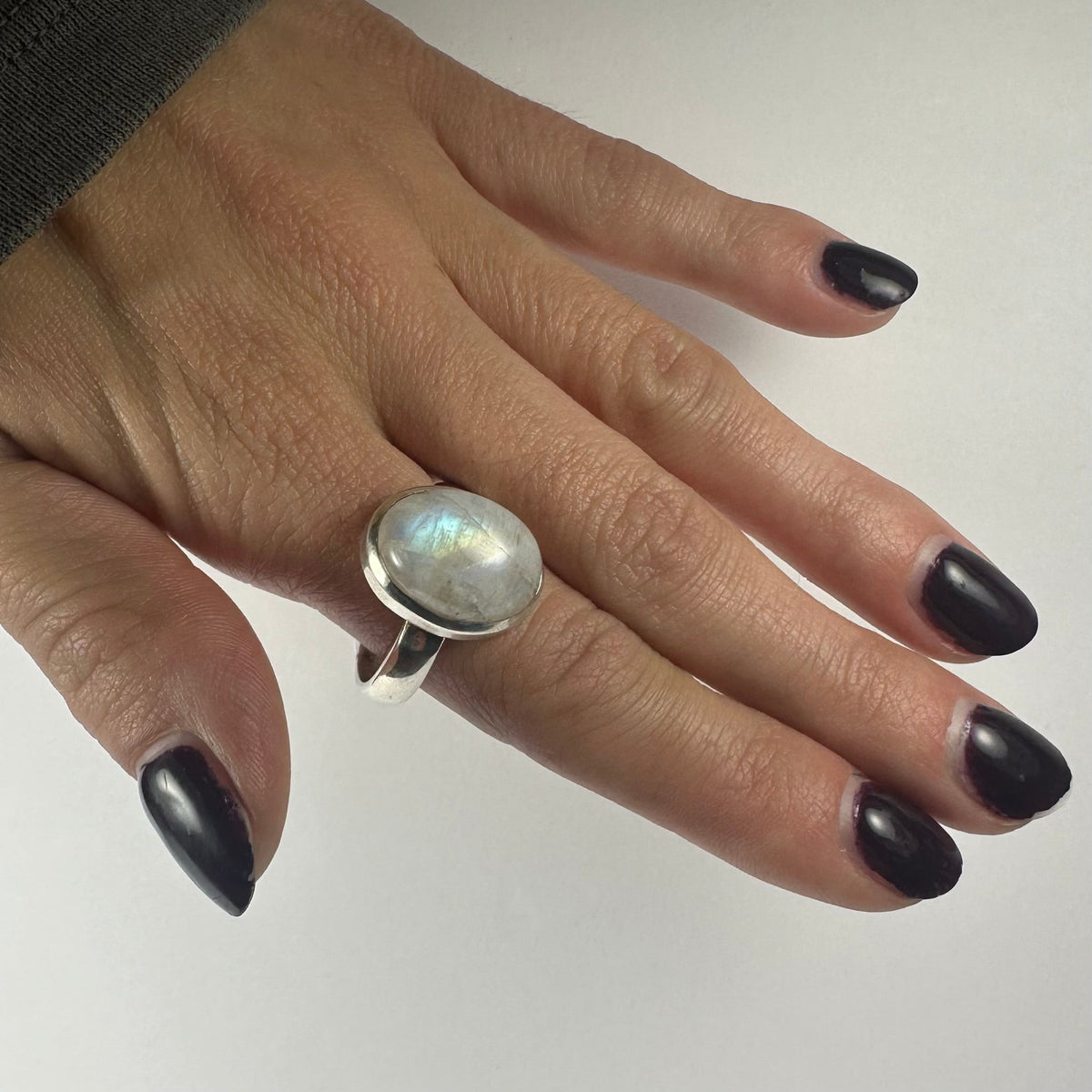 Hand with dark polished nails wearing a silver ring featuring a round, iridescent moonstone, shown against a plain light background.