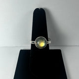 Silver ring with a round labradorite stone showing a subtle yellow and green flash, displayed on a black velvet stand against a plain background.