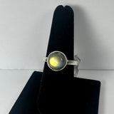 Silver band ring with a round labradorite stone showing yellow and green iridescence, displayed on a black velvet finger stand against a white background.