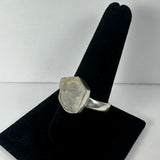 Silver ring with a large, rough clear quartz stone set in an irregular hexagonal bezel on black velvet ring display stand