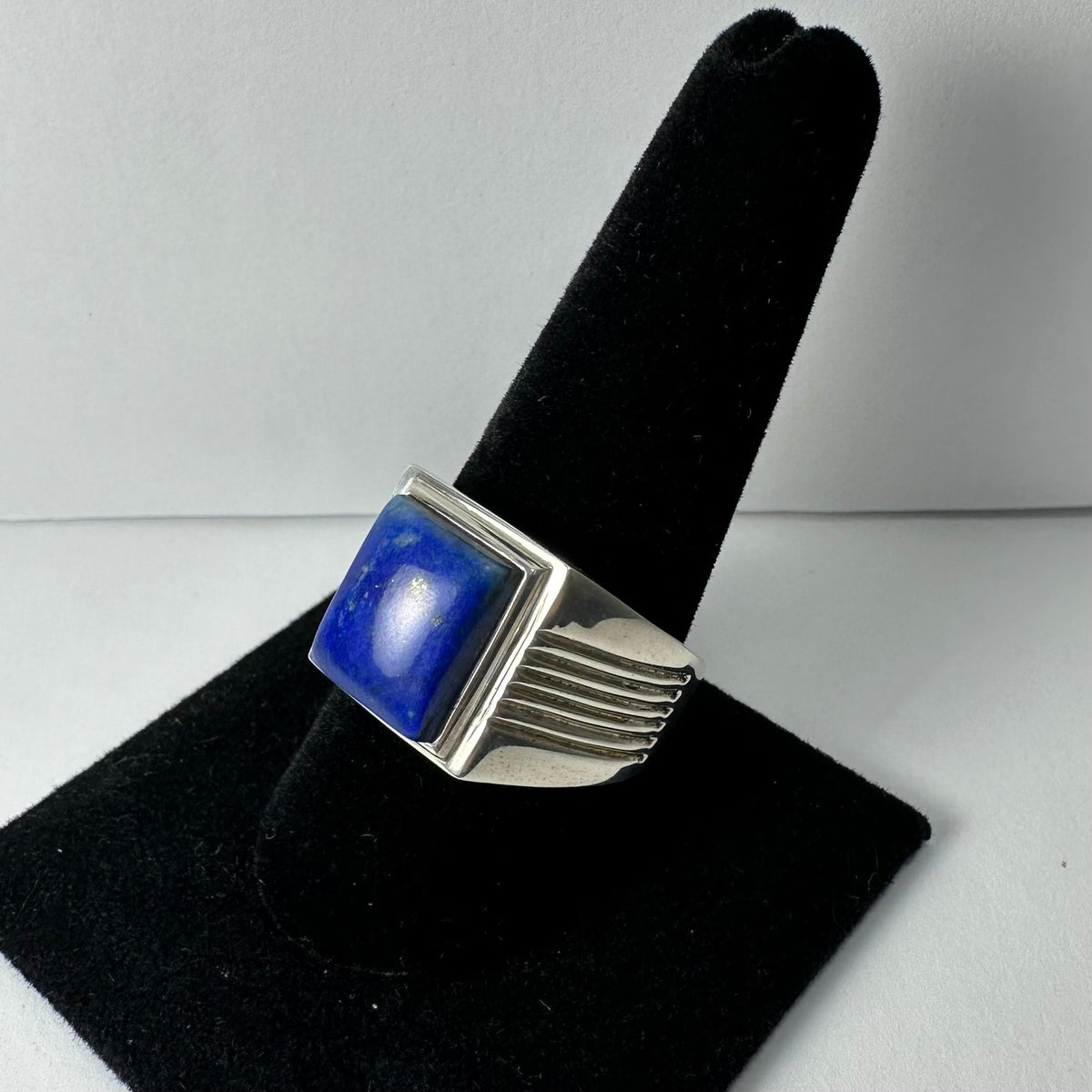 Silver ring with a polished square lapis lazuli stone set in a simple bezel, displayed on a black velvet ring stand showing a side view with ribbed band details.