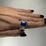 Hand with dark purple nails wearing a silver ring featuring a rectangular deep blue lapis lazuli stone and parallel engraved lines on the band.