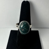 Silver ring with a polished oval green stone featuring white and gray inclusions, displayed on a black velvet ring holder against a plain background.