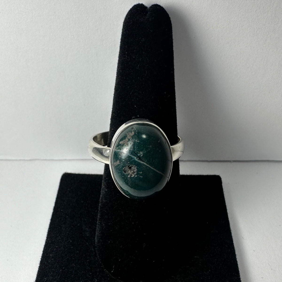 Silver ring with a polished oval green stone featuring white and gray inclusions, displayed on a black velvet ring holder against a plain background.