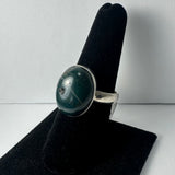 Silver ring with a polished oval green serpentine stone featuring natural darker inclusions, displayed on a black velvet finger stand against a white background.