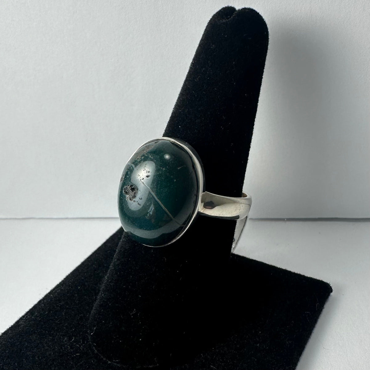 Silver ring with a polished oval green serpentine stone featuring natural darker inclusions, displayed on a black velvet finger stand against a white background.