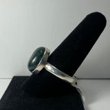 Silver ring with a dark green, glossy cabochon stone displayed on a black velvet finger stand against a plain gray background.