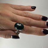 Hand wearing a silver ring with a polished dark green serpentine stone, with black and white nail polish on a plain white background.