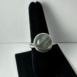 Silver ring with a round, translucent labradorite stone displaying gray and green hues, shown on a black velvet ring holder against a plain background.