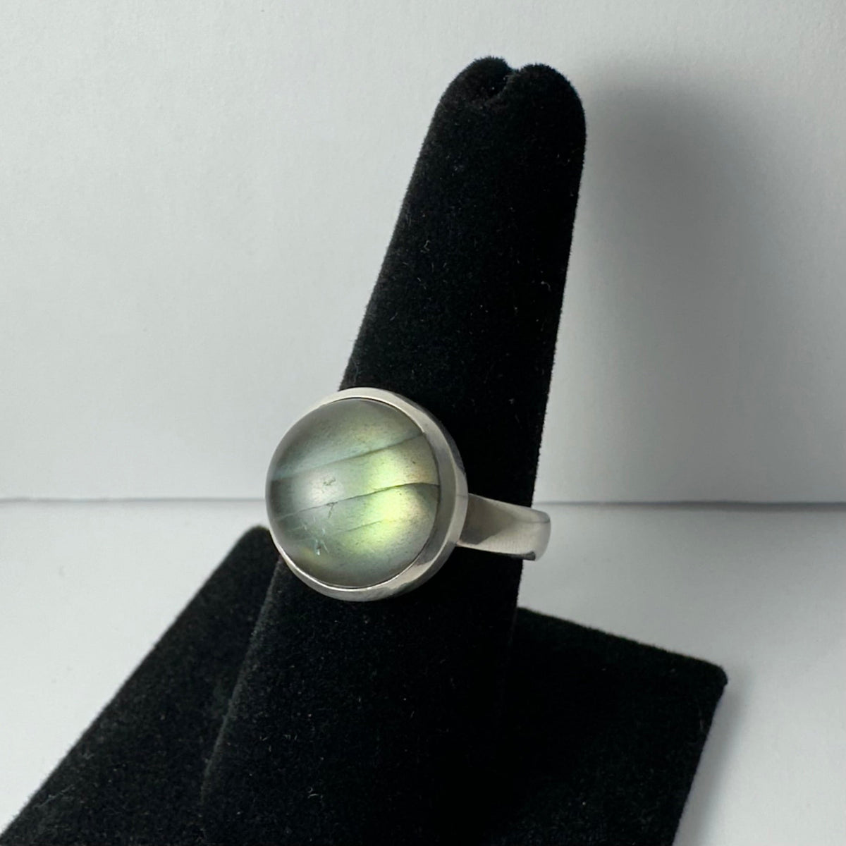 Round labradorite ring with a smooth polished stone showing green and blue iridescence, set in a simple silver band on a black velvet finger stand.