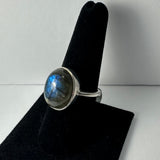 Silver ring with a round, polished labradorite stone displaying blue and gray hues, shown on a black velvet ring holder against a white background.