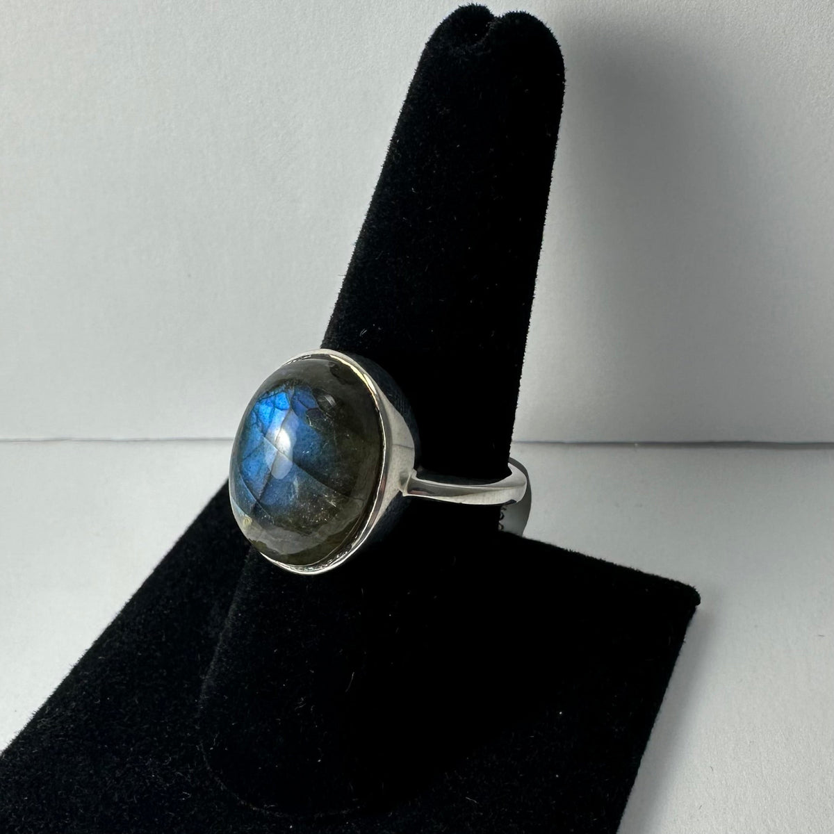 Silver ring with a round, polished labradorite stone displaying blue and gray hues, shown on a black velvet ring holder against a white background.