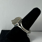 Silver ring with a raw, irregular clear quartz crystal set on a double band, displayed on a black velvet ring holder against a gray background.