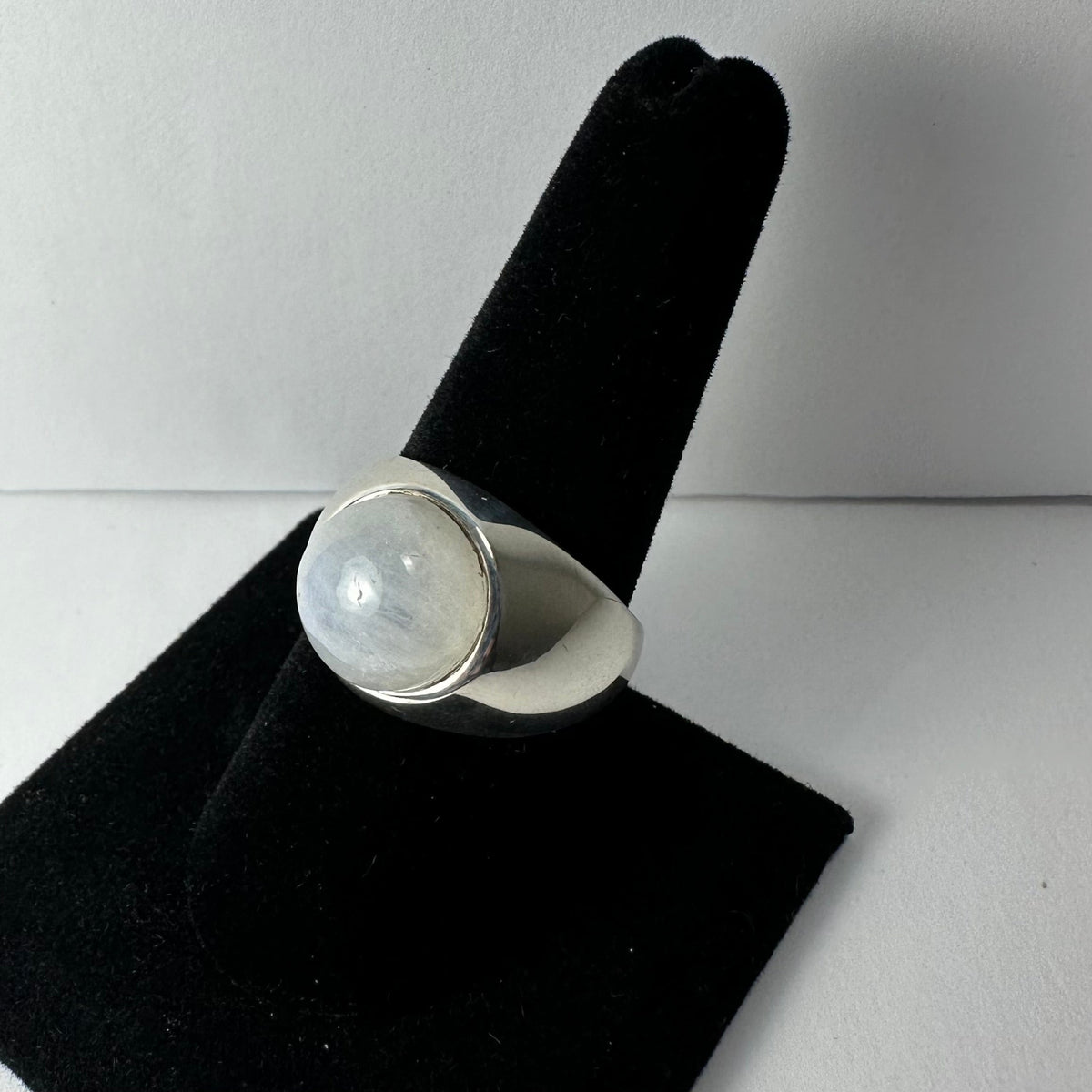 Silver ring with a round, polished moonstone set in a smooth bezel, displayed on a black velvet finger-shaped stand against a pale background.