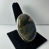 Oval polished labradorite stone with metallic gray and gold hues set in a silver-colored metal ring, displayed on a black velvet ring stand against a white background.