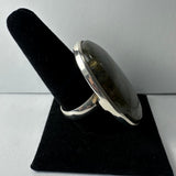 Large oval labradorite stone ring with a smooth silver bezel and band displayed on a black velvety ring holder against a gray background.