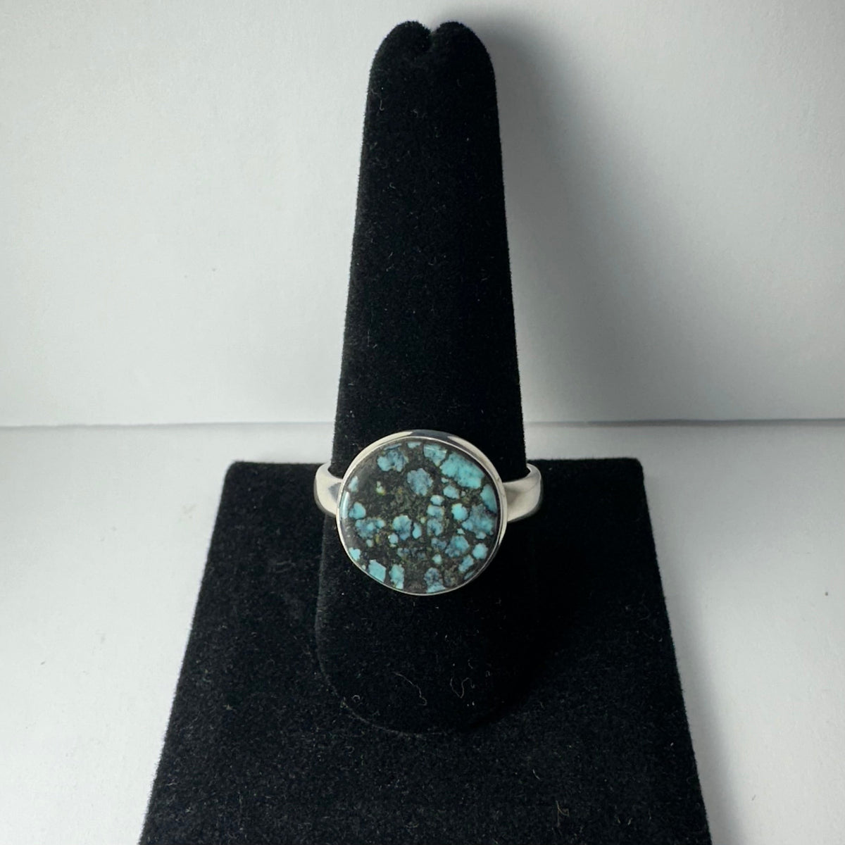 Round Tibetan turquoise ring with a silver band and a mottled blue and black stone set in a circular bezel, displayed on a black velvet holder.